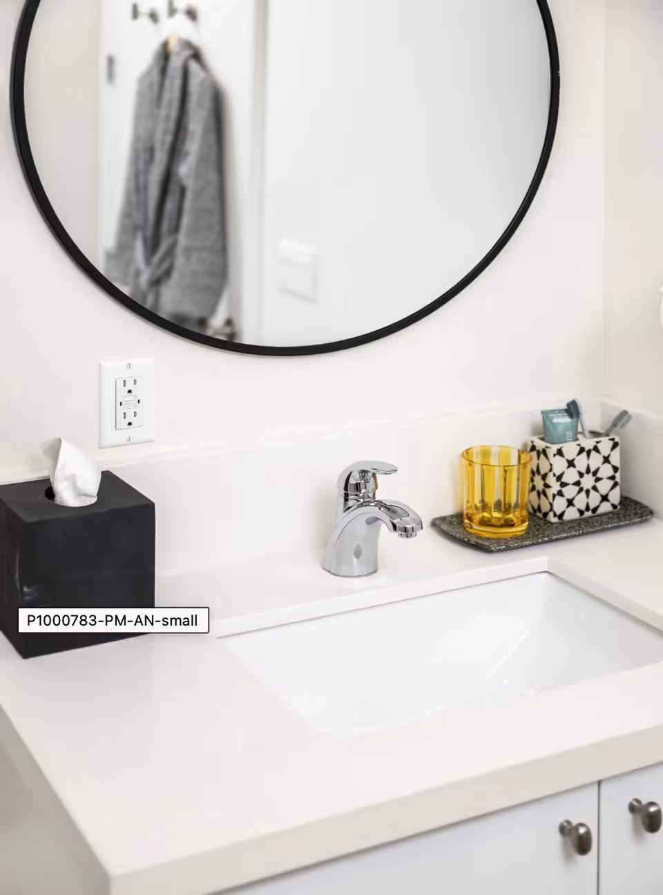 A bathroom sink area with a chrome faucet, a black tissue box, a yellow glass, and a black and white patterned container holding toothbrushes and toothpaste. A round mirror with a black frame hangs above the sink, reflecting a gray robe hanging on a hook on the door.