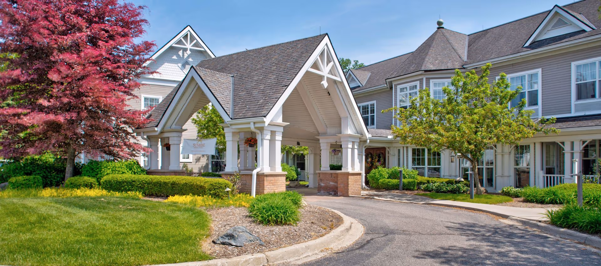 Front entrance and porte-cochère of a senior living building with landscaped lawns and a curved driveway.