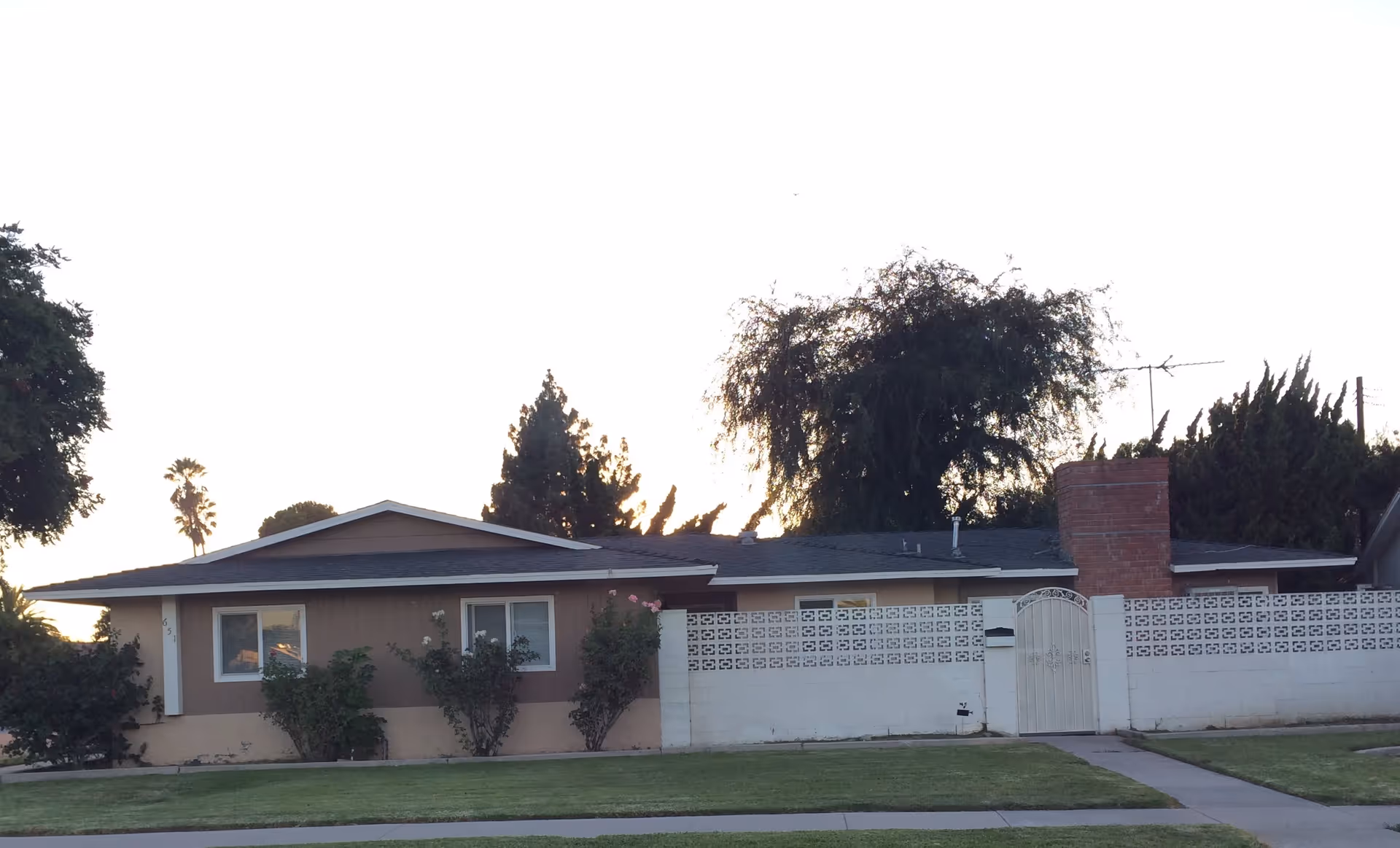 Single-story residential house with a low decorative block wall and gated entry, a front lawn and trees in front at sunset.