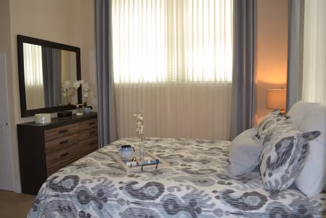 A neatly made bed with patterned bedding and pillows in a softly lit bedroom, with a dresser and mirror to the left and a curtained window behind.