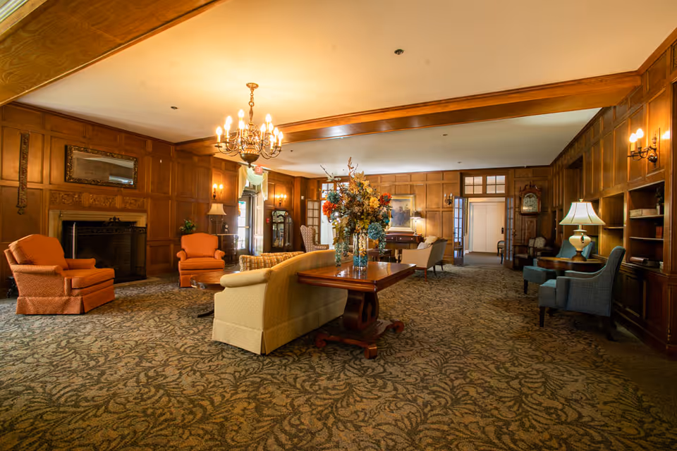A spacious senior living community lounge with wood-paneled walls, patterned carpet, and a chandelier. The room features comfortable seating including orange armchairs, a beige sofa, and blue chairs. There is a wooden table with a large floral arrangement in the center, a fireplace with a mirror above it, and various lamps providing warm lighting.