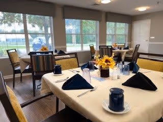 A dining room with tables covered in white tablecloths, each set with blue napkins, blue cups, and a centerpiece of yellow flowers. Large windows with blinds allow natural light to fill the room, and chairs are arranged around the tables.