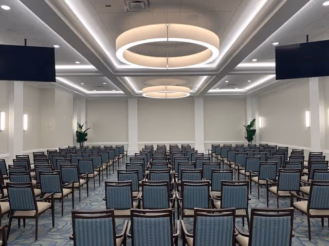 Large, empty conference/event room with rows of blue-upholstered chairs facing a blank front wall under circular ceiling lights.