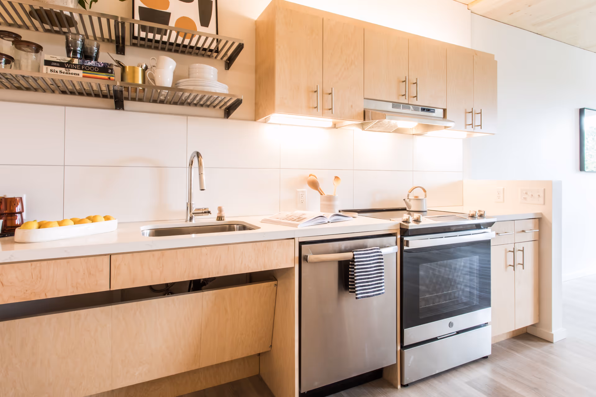 Modern kitchen with light wood cabinets, a stainless steel dishwasher and oven, a sink with a chrome faucet, open shelves holding dishes and books, and a countertop with a tray of lemons and wooden utensils.