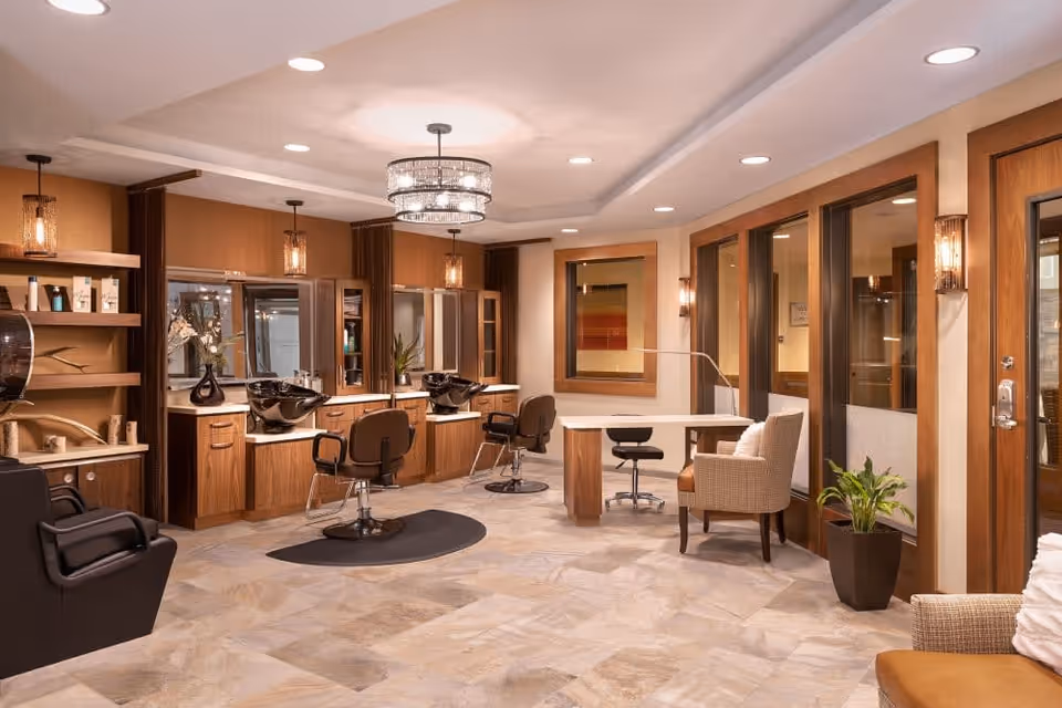 Well-lit salon area with styling stations, wash sinks, chairs, a manicure table, and cozy seating in warm wood tones.