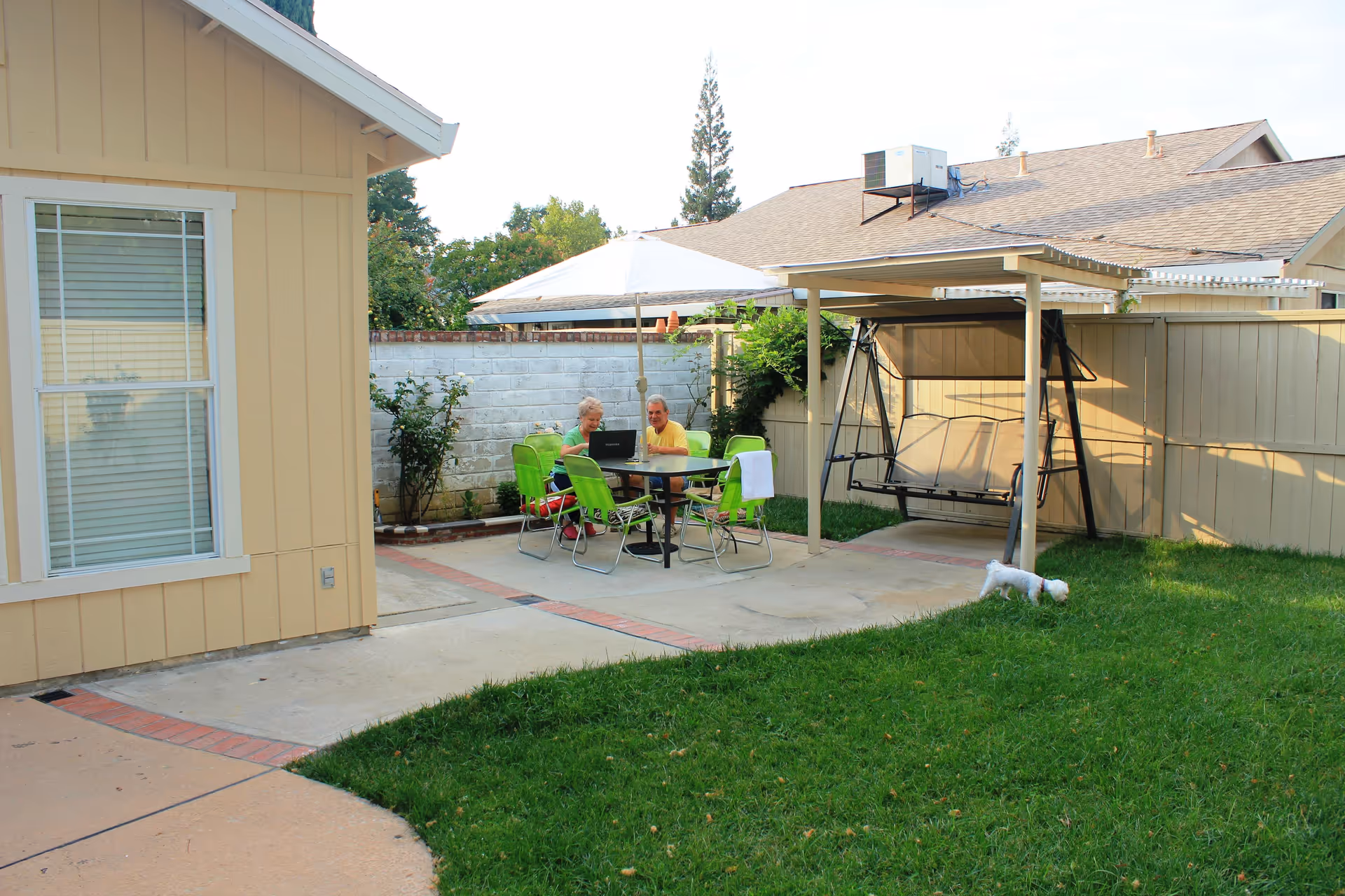 An outdoor patio area with a table and green chairs under a white umbrella. Two elderly people are sitting at the table using a laptop. There is a small white dog on the grass nearby and a covered swing seat next to a beige fence. The scene is part of a residential facility with a yellow building visible on the left.