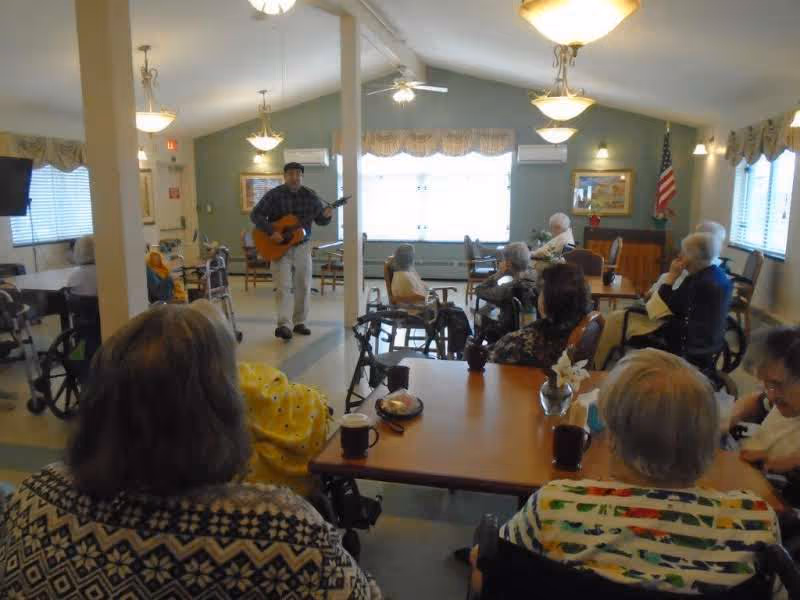 A group of elderly people seated around tables in a common room, watching a man playing a guitar. The room is well-lit with ceiling lights and has windows with curtains. Some people are in wheelchairs, and there is an American flag in the corner.