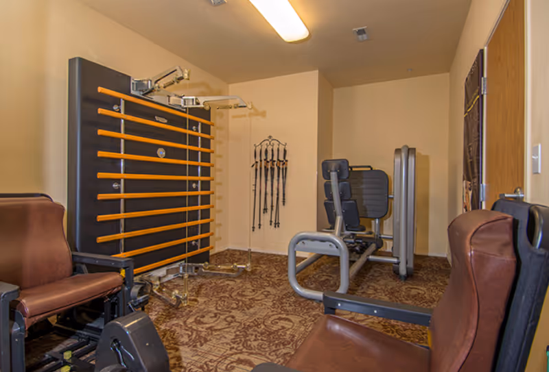 A small exercise room with padded brown chairs, a wall-mounted exercise machine with wooden bars, a leg press machine, and several walking poles hanging on the wall. The room has beige walls and patterned carpet flooring.