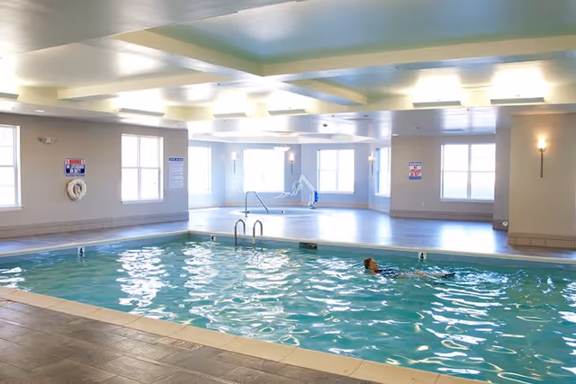 Indoor swimming pool with a person swimming, pool lift, large windows, and recessed ceiling lighting.