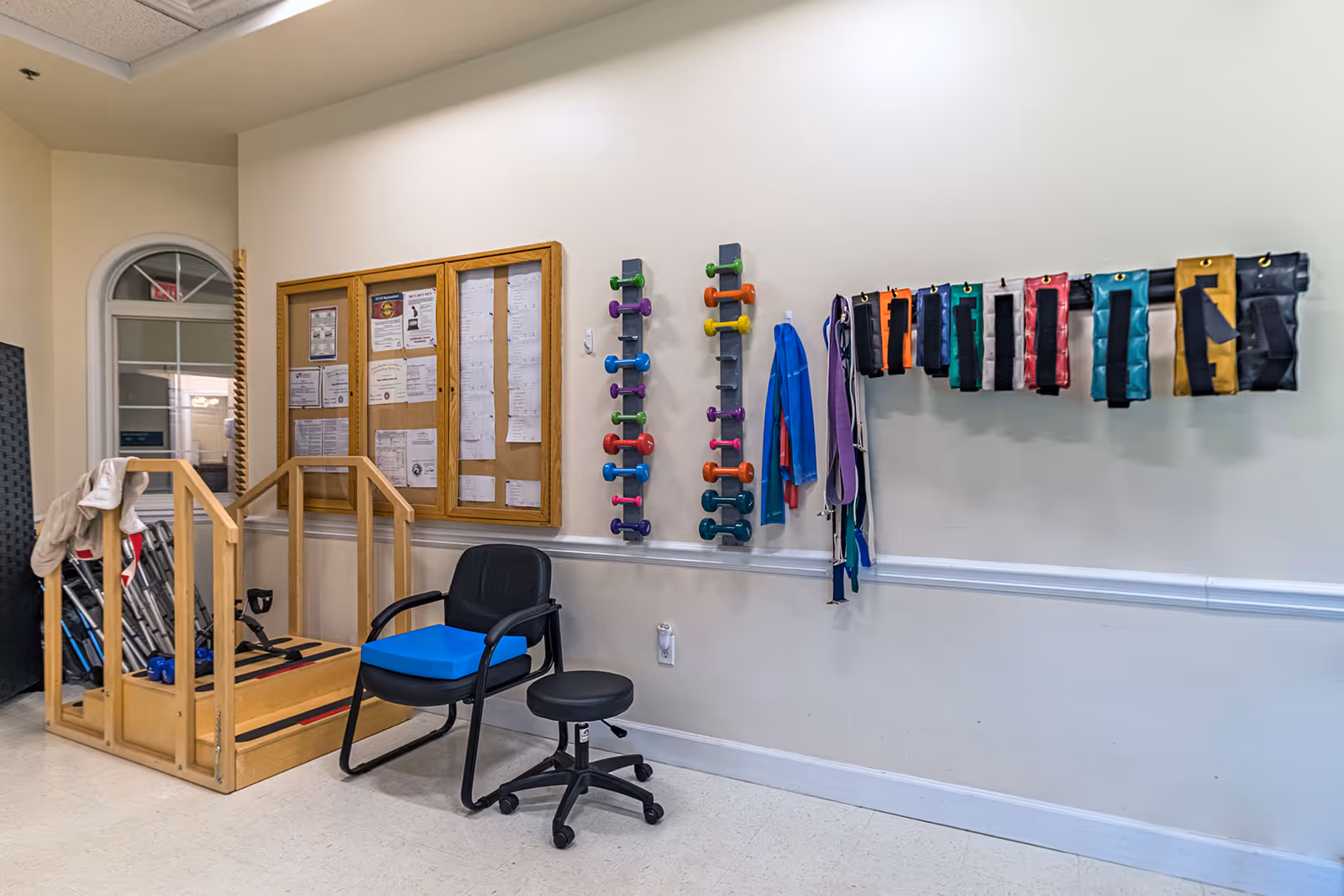 Interior view of a senior living facility exercise area with a wooden ramp, a black chair with a blue cushion, a black rolling stool, colorful dumbbells mounted on the wall, resistance bands hanging on hooks, and a bulletin board with notices.
