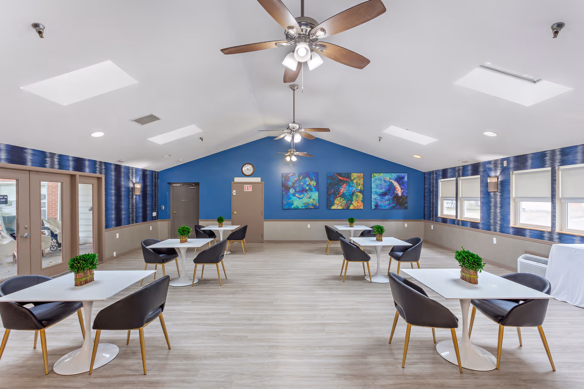 Bright communal dining/activity room with round white tables, modern chairs, ceiling fans, and colorful artwork on a blue accent wall.