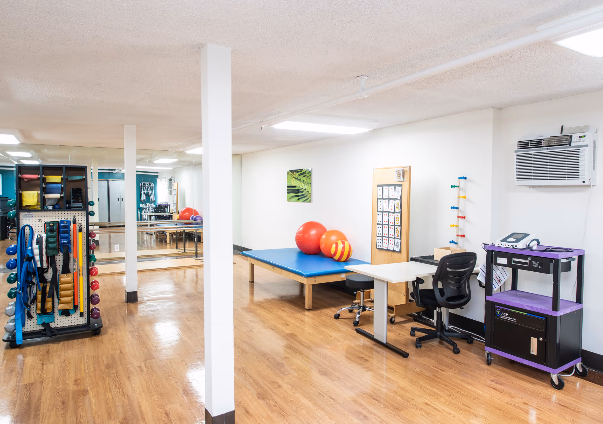 A physical therapy or rehabilitation room with exercise equipment including resistance bands, weights, therapy balls, and a padded therapy table. There is a desk with a chair, a corkboard with cards, and an air conditioning unit mounted on the wall. The room has wooden flooring and mirrored walls.