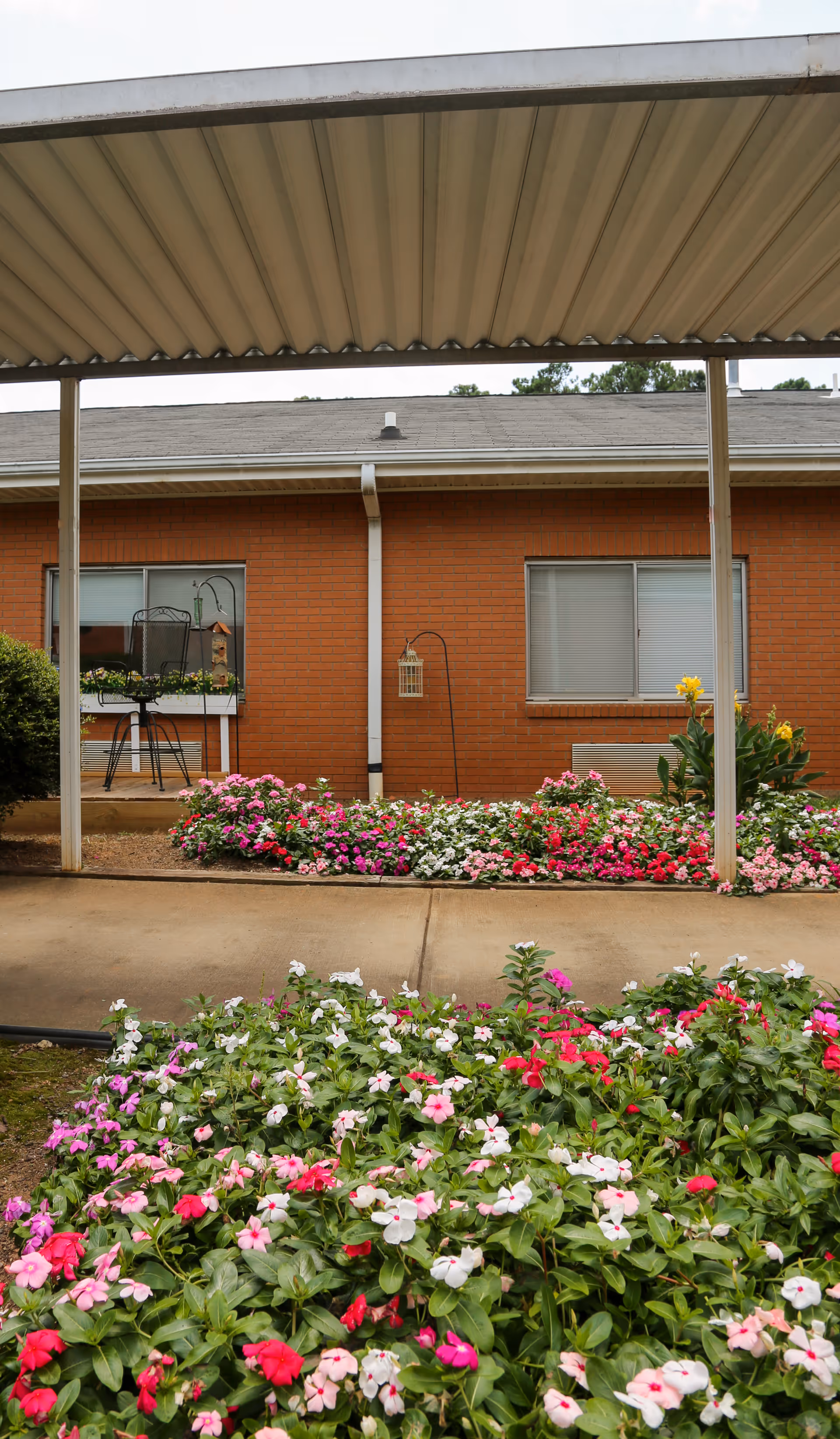 Covered walkway with colorful flower beds on both sides in front of a brick building with windows and a small patio area with a chair and bird feeder.