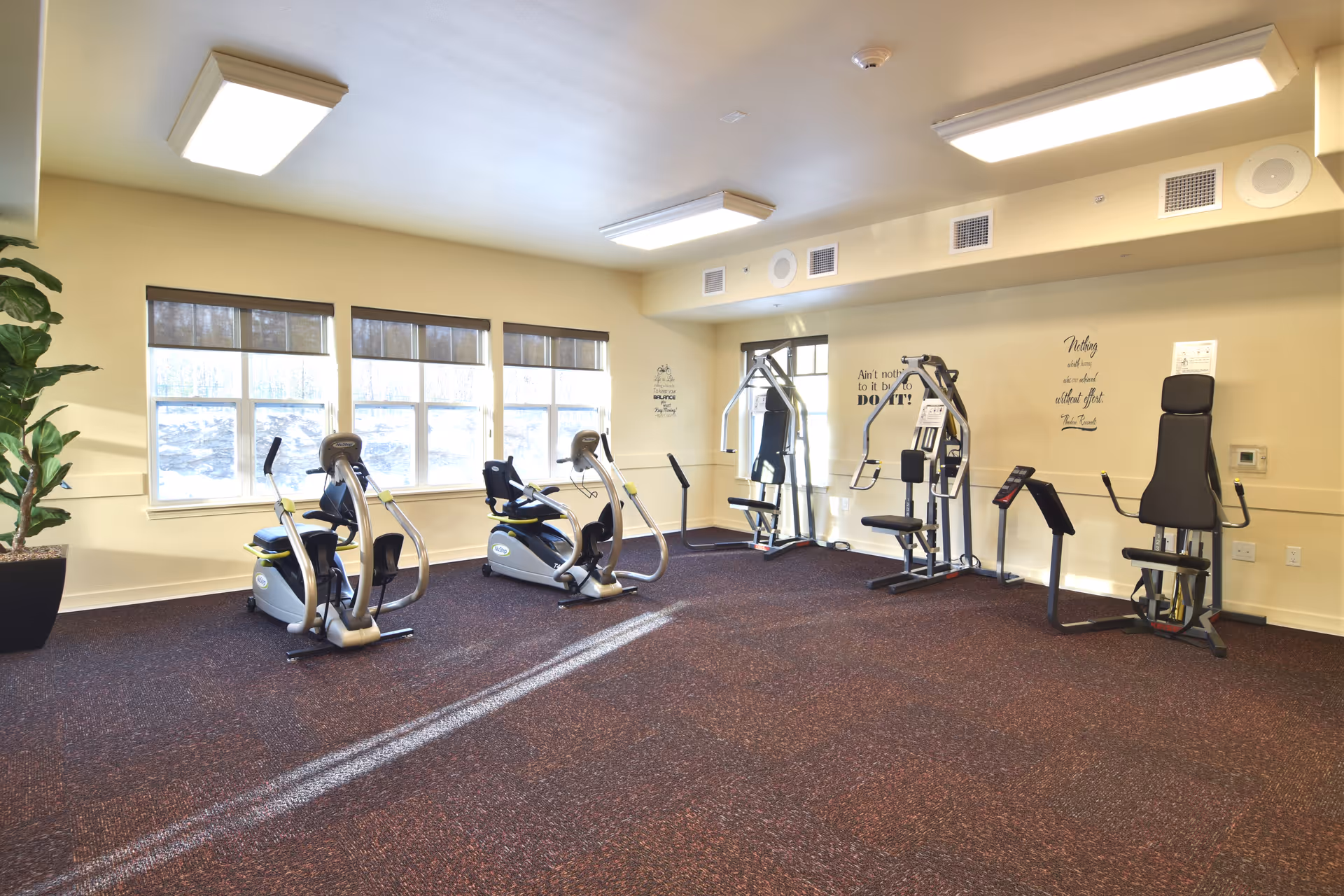 A bright exercise room with several pieces of fitness equipment including two recumbent bikes, a multi-function weight machine, and other exercise machines. The room has large windows letting in natural light, beige walls with motivational quotes, and a dark carpeted floor.