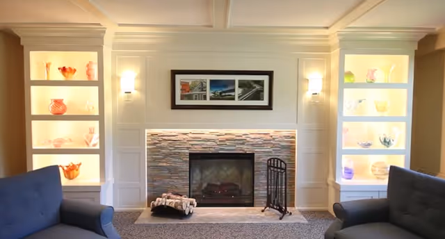 A cozy living room featuring a stone fireplace with a black metal screen and a log holder filled with firewood. On either side of the fireplace are built-in shelves illuminated with warm lighting, displaying decorative glass vases and bowls. Two blue upholstered armchairs face the fireplace, and above it hangs a framed triptych photograph of architectural scenes. The room has a soft carpet and paneled walls with sconces providing additional light.