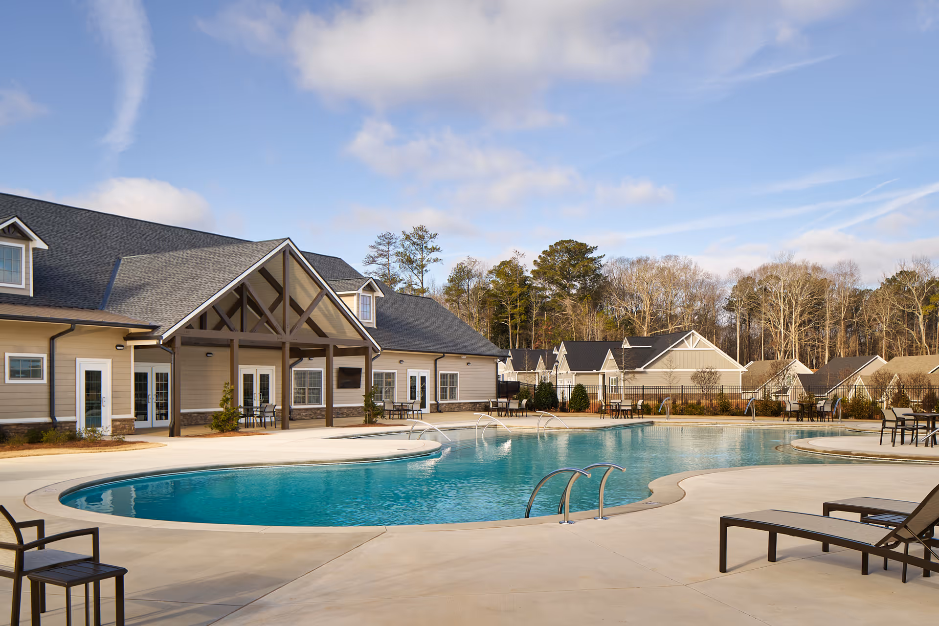 Outdoor swimming pool area with lounge chairs and tables, adjacent to a building with a covered patio. Trees and additional buildings are visible in the background under a partly cloudy sky.