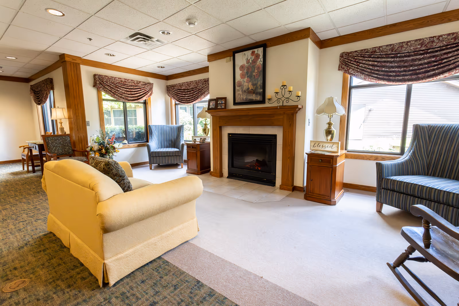 A cozy living room area in an assisted living facility featuring a beige sofa, two blue striped armchairs, a wooden rocking chair, and a fireplace with a floral painting above it. The room has large windows with patterned valances allowing natural light to fill the space. There are wooden side tables with lamps and decorative items, including a sign that says 'blessed'.