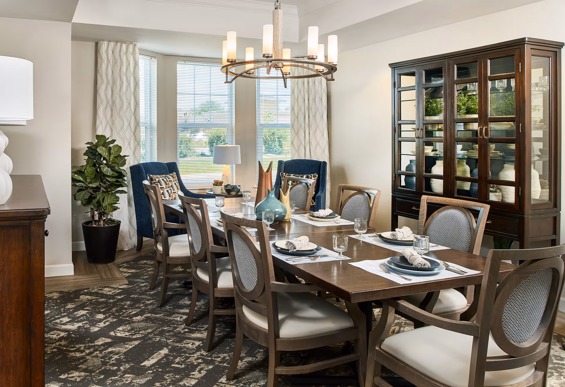 A well-lit dining room with a long wooden table set for eight people, featuring plates, glasses, and napkins. The room has large windows with white curtains, two blue upholstered chairs at the far end, a dark wooden china cabinet filled with decorative items, a potted plant, and a modern chandelier hanging above the table.
