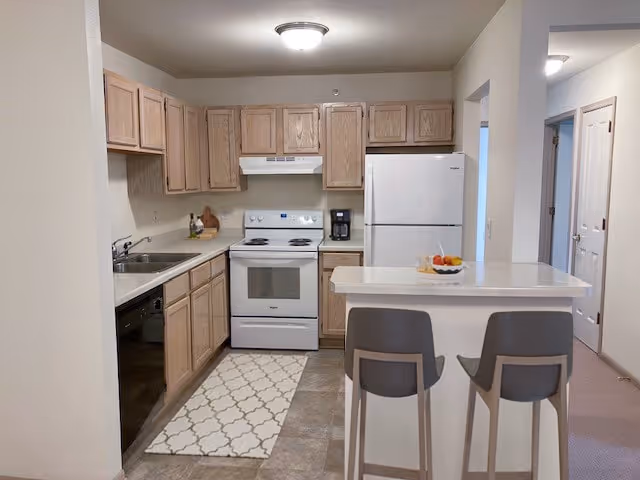 A compact kitchen in The Silvernail Senior Apartments featuring light wood cabinets, a white electric stove with oven, a white refrigerator, a dishwasher, and a coffee maker. There is a small island with two gray bar stools and a bowl of fruit on top. The floor has a patterned rug in front of the stove and tile flooring near the island. The walls are painted white, and there is a ceiling light fixture.