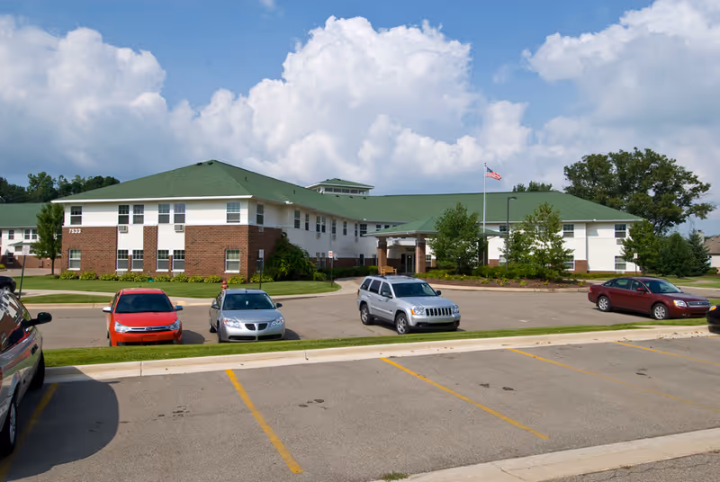 Exterior view of Woodland Village senior living facility showing a two-story building with a green roof and brick and white siding. Several cars are parked in the parking lot in front of the building under a partly cloudy sky.