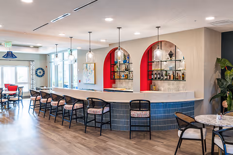 A modern indoor bar area with a curved white marble countertop and blue tiled base. There are six black bar stools with patterned cushions lined up along the bar. Behind the bar are two arched shelves with red back panels displaying various bottles and glassware. The room has wooden flooring, pendant lights hanging above the bar, and additional seating with tables and chairs in the background near large windows.