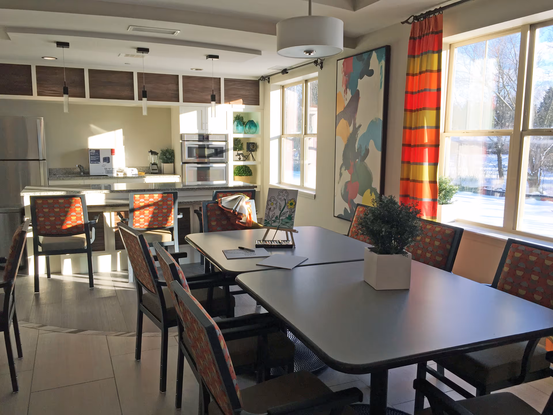 Bright communal dining area and kitchen with tables, patterned chairs, pendant lights, and large windows.