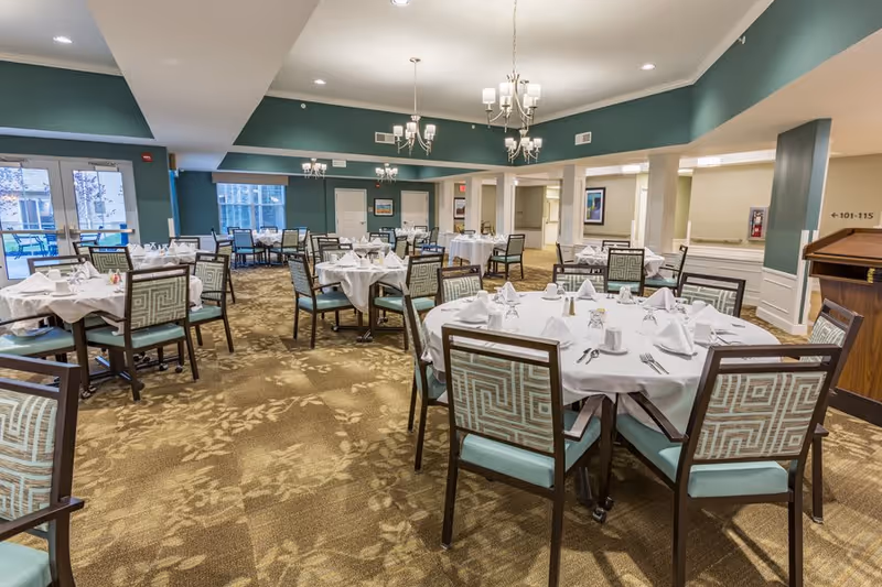 A spacious dining room with multiple round tables covered with white tablecloths, each set with folded napkins, glasses, and silverware. The room features patterned carpet, teal walls with white trim, and several chandeliers hanging from the ceiling. Large windows and glass doors allow natural light to enter the room.
