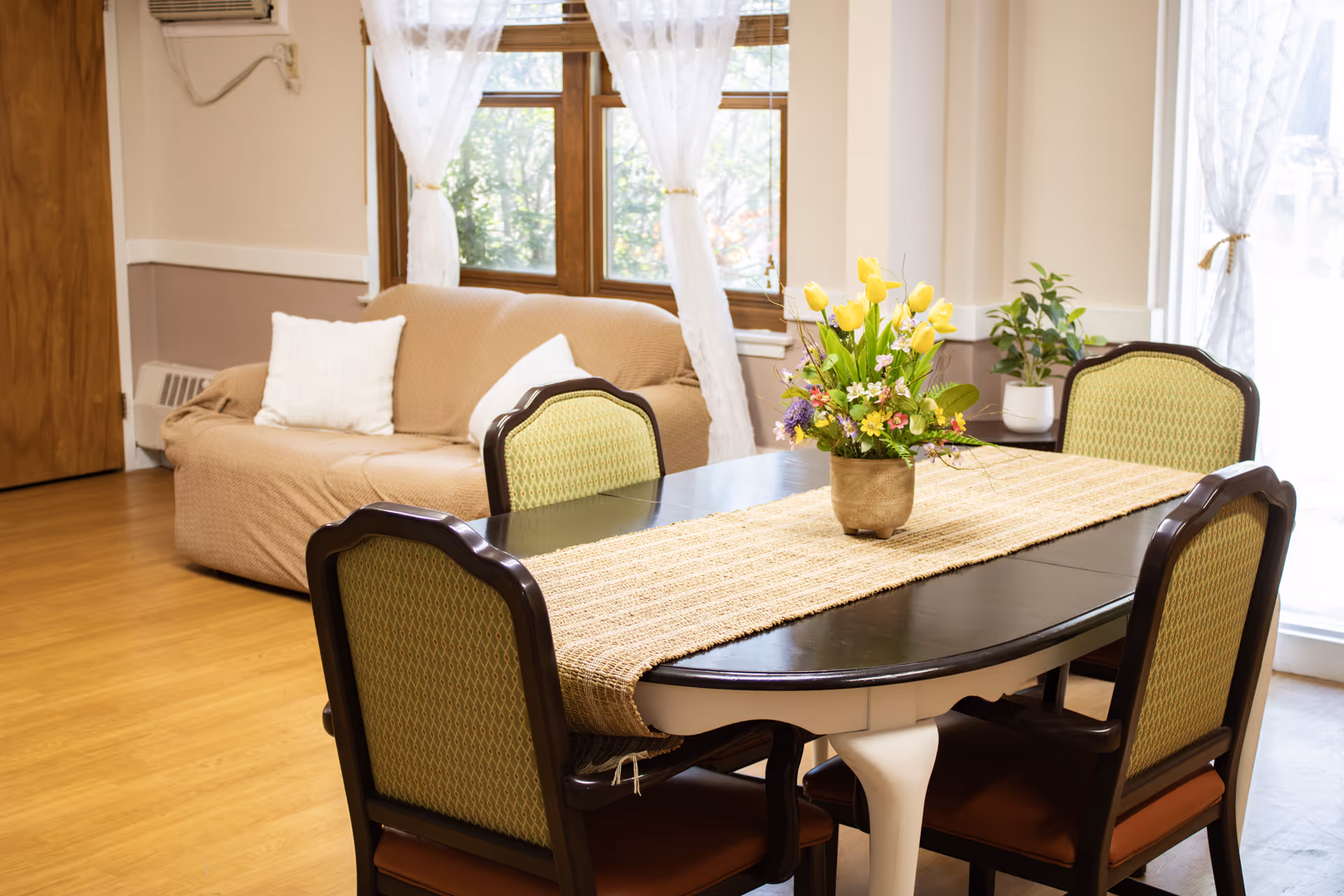 Dining table with four upholstered chairs and a vase of yellow flowers in a bright common room with a sofa and windows.