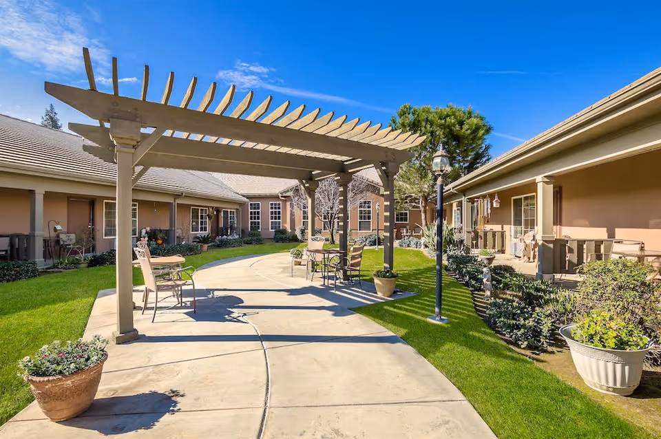 Outdoor courtyard area at a senior living facility with a pergola providing partial shade over a concrete pathway. There are several chairs and tables arranged under the pergola and along the pathway. The courtyard is surrounded by single-story buildings with windows and doors, and there are potted plants and greenery along the edges. A lamppost is also visible near the pergola.