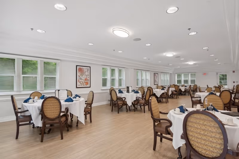 A spacious dining room with multiple round tables covered in white tablecloths, each set with blue napkins, cups, and menus. The room has large windows with white blinds, light wood flooring, and framed artwork on the walls. The chairs have wooden frames with patterned upholstery.