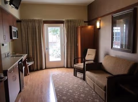 Small studio living area with a couch and armchair, a kitchenette on the left, and a glass door with curtains leading outside.