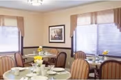 A dining room with round tables set for a meal, featuring white tablecloths, cups, plates, and yellow flower centerpieces. The room has beige walls, two windows with blinds and valances, and a framed picture on the wall.