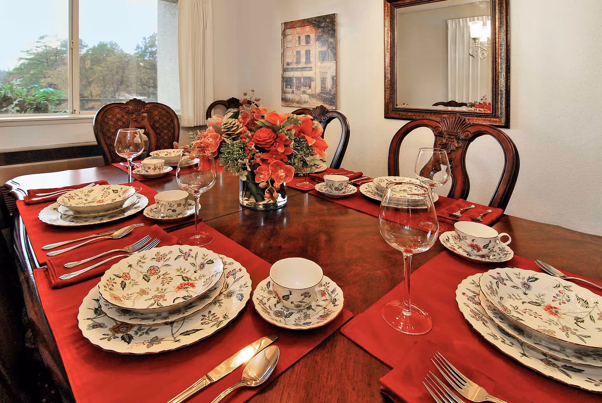 A formal dining table set with floral-patterned plates, bowls, and teacups on red placemats. The table has ornate wooden chairs, a centerpiece with red and pink flowers, and wine glasses. A large window with a view of trees and a framed painting and mirror are visible on the walls.