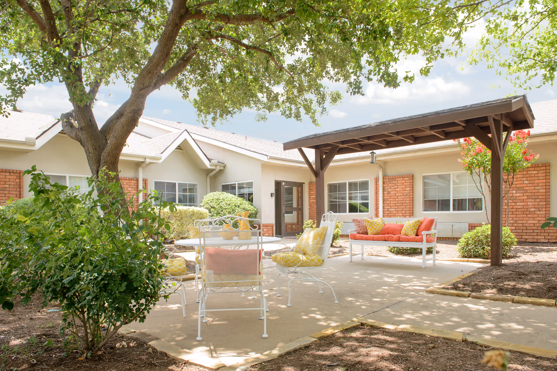 Shaded courtyard patio at a senior living facility with white metal chairs, a cushioned bench under a wooden pergola, and surrounding brick-faced building.