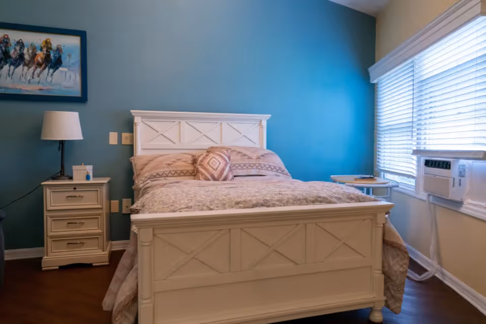 Neat bedroom with a white wooden bed and nightstand against a teal accent wall, a lamp, framed art, and a window with blinds and an air conditioner.