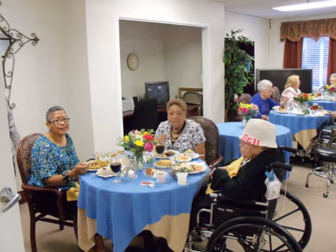 Several elderly residents sit at round tables with blue tablecloths and flowers while eating a meal in a communal dining room.