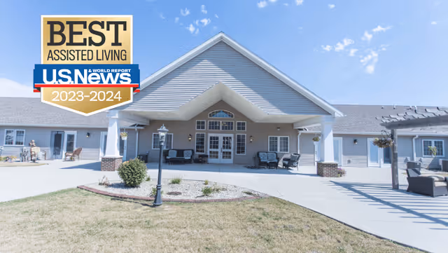 Exterior view of Villas of Holly Brook Newton, a single-story assisted living facility with a large covered entrance, outdoor seating areas, and a clear blue sky. There is a landscaped area with a lamp post in front of the building.