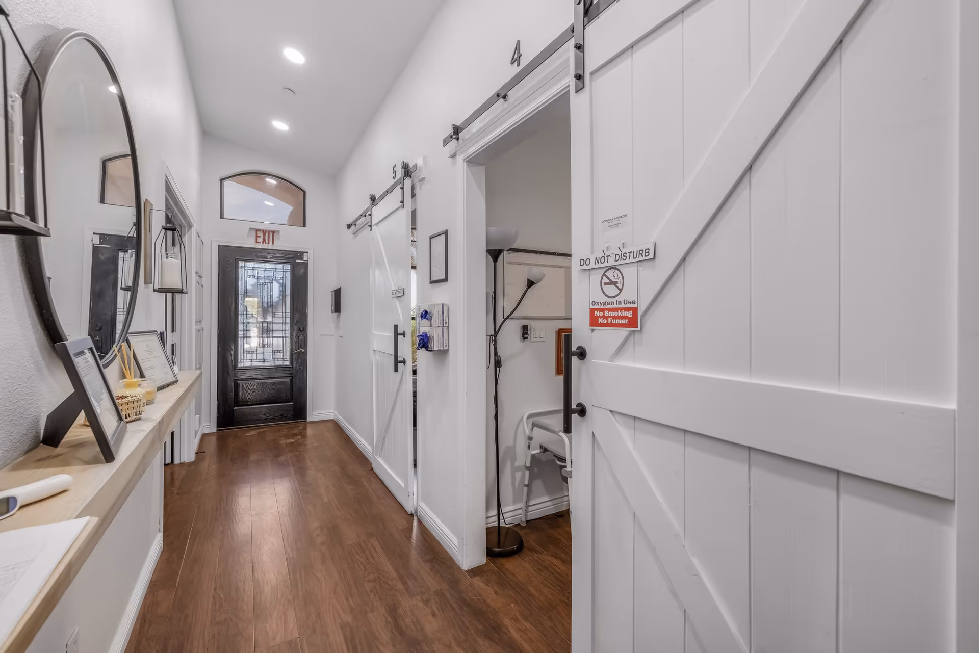 A clean, well-lit hallway with wooden flooring and white walls. On the right side, there are white sliding barn doors labeled with numbers, one of which is partially open showing a room with a chair and a floor lamp. A 'Do Not Disturb' sign with warnings about oxygen use and no smoking is hanging on the open door. On the left side, there is a narrow shelf with framed pictures, a mirror, and some decorative items. At the end of the hallway, there is a black door with a glass panel and an exit sign above it.