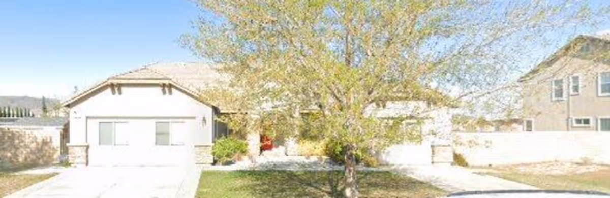 Front exterior view of a single-story residential building with a driveway, a tree in the front yard, and a clear blue sky.