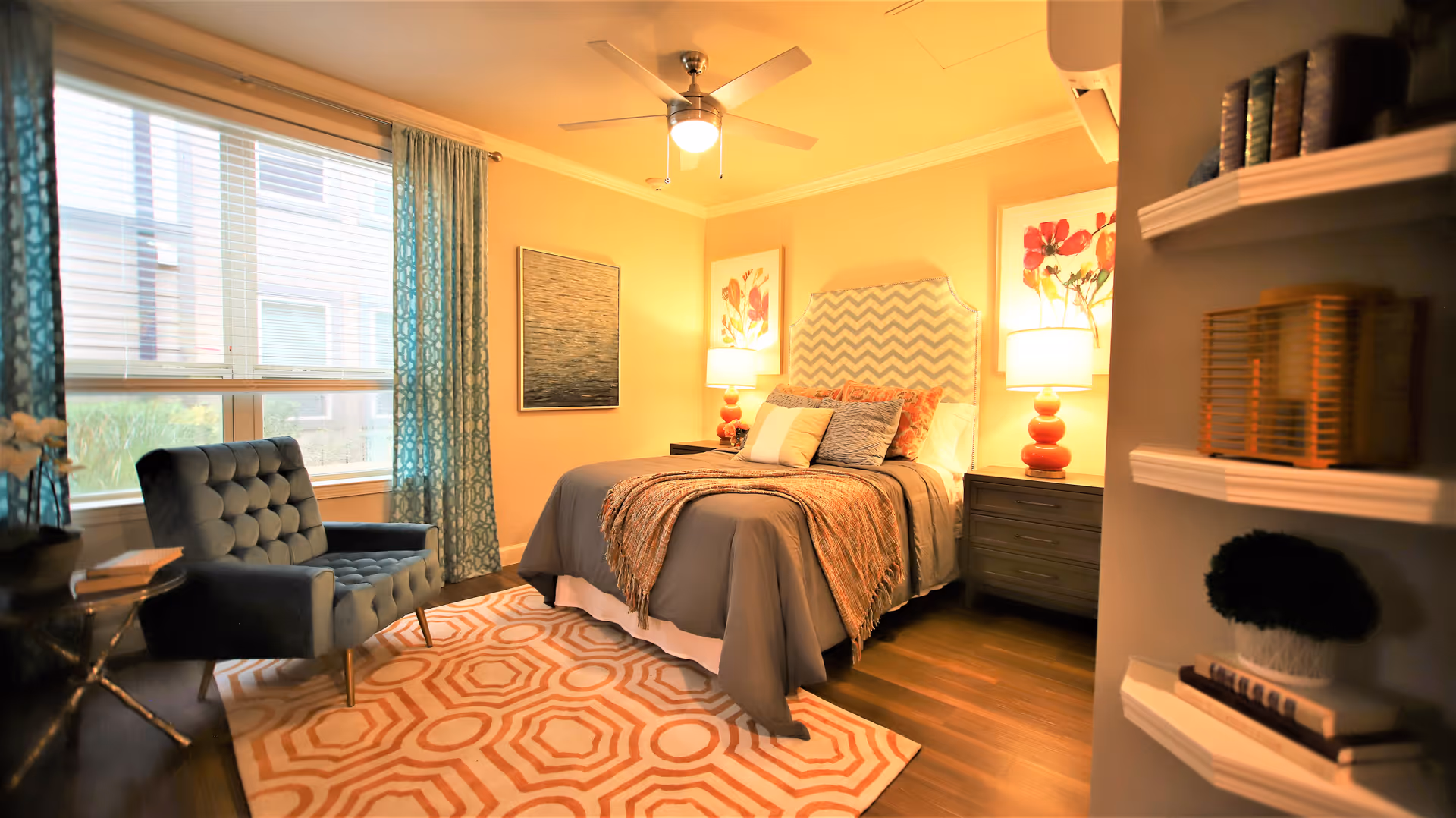 Well-lit, warmly decorated bedroom with a bed flanked by bedside lamps, a patterned rug, and an upholstered armchair by the window.