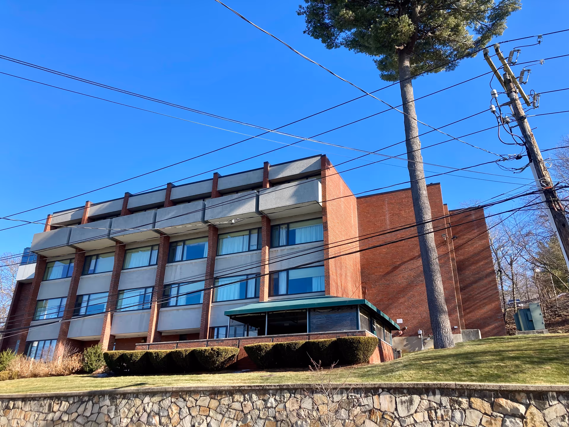 Exterior view of a multi-story brick and concrete building with large windows, a green roofed entrance area, and a stone retaining wall in front. There are power lines and a tall tree visible against a clear blue sky.