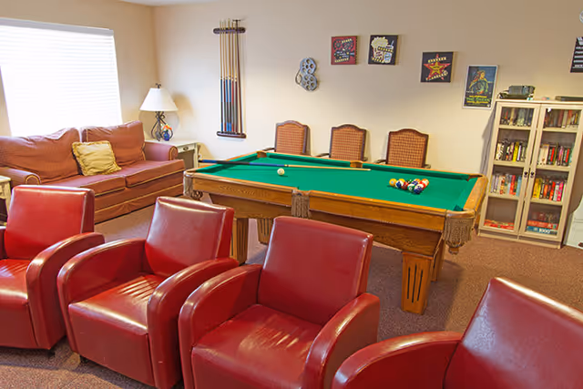 A cozy community recreation room with a pool table, red leather chairs, a sofa, and a bookshelf.