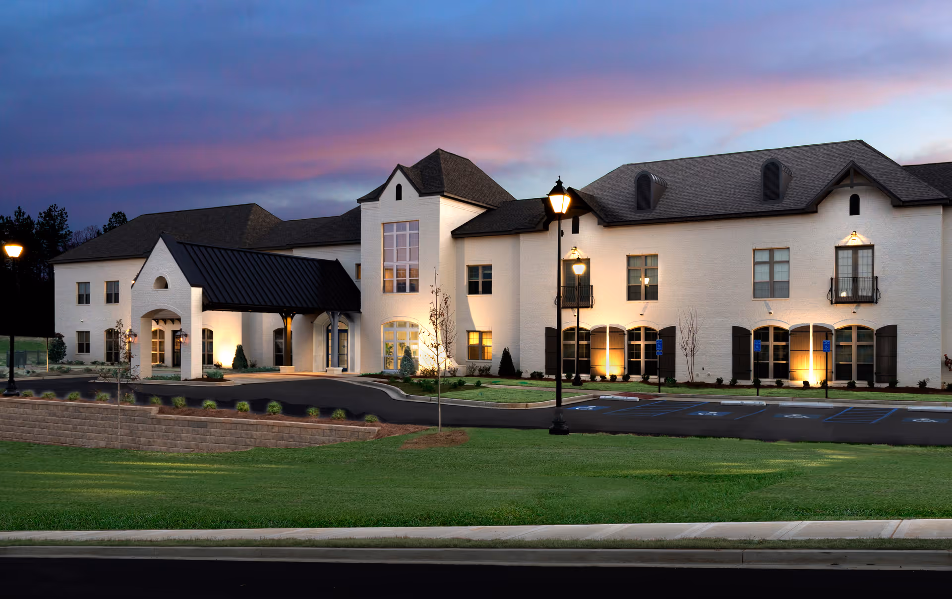 Exterior view of a large, two-story senior living facility building at dusk with white brick walls, black roof, and several lit windows. The entrance has a covered drop-off area with black metal roofing. There are street lamps and a parking lot with marked handicapped spaces in front of the building, surrounded by well-maintained grass and landscaping.