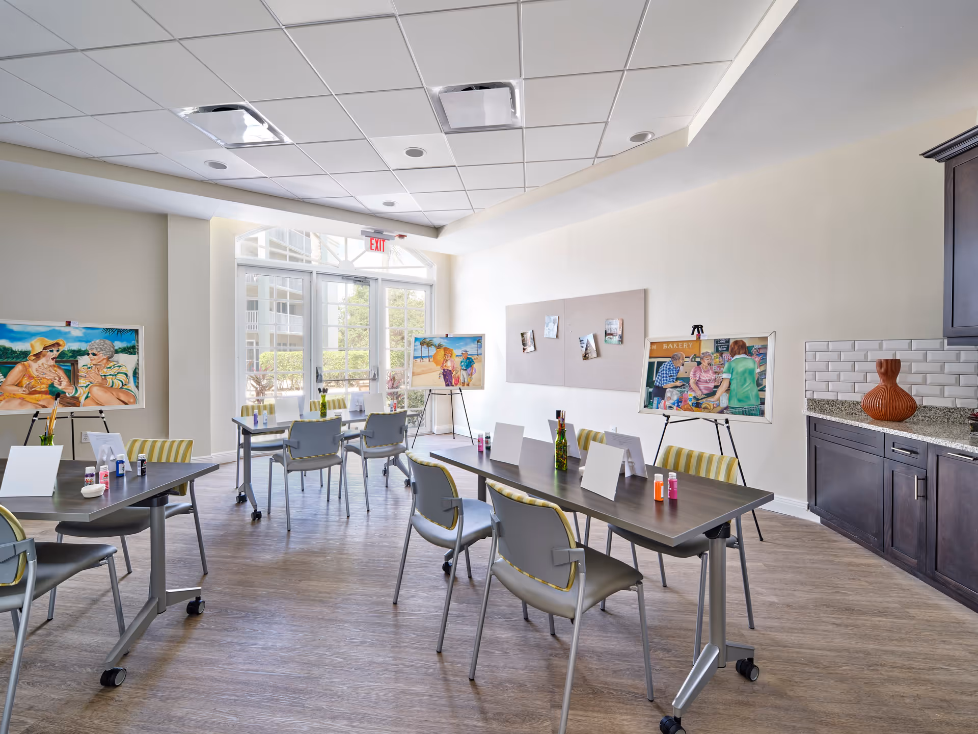 A bright, spacious room with tables and chairs arranged for an art activity. Paint bottles, brushes, and blank canvases are set up on the tables. Colorful paintings on easels and a bulletin board with pinned photos decorate the room. Large windows let in natural light, and there is a countertop with cabinets on the right side.