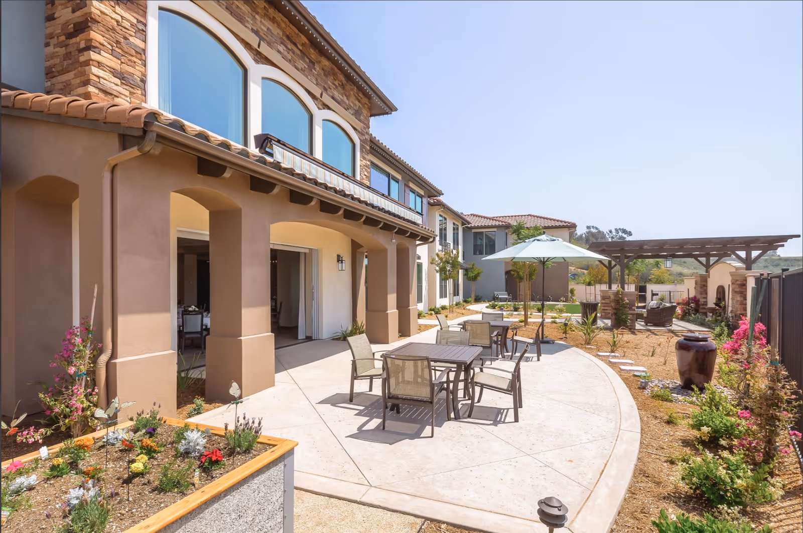 Outdoor courtyard with patio dining tables and umbrella, landscaped paths, and a multi-story building in the background.