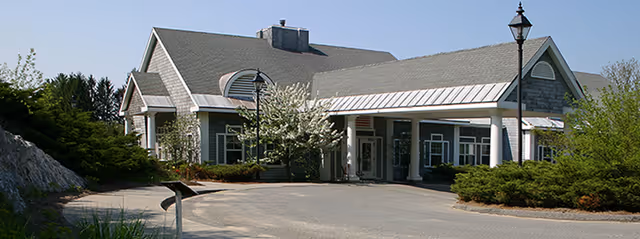 Exterior view of Sharon Health Care Center showing a single-story building with a gray roof, green siding, white trim, and a covered entrance. There are bushes and trees around the building and a lamppost near the driveway.