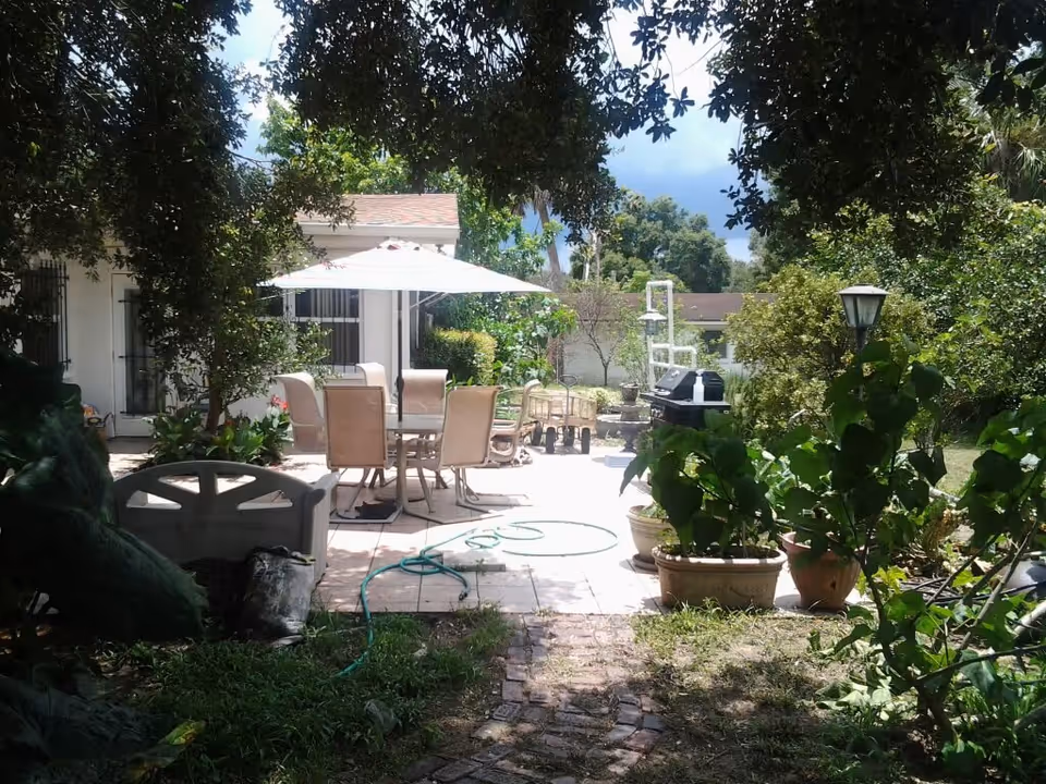 Outdoor patio area with a table and six chairs under a large white umbrella. The patio is surrounded by greenery including potted plants and trees, with a garden hose on the tiled floor and a barbecue grill in the background.