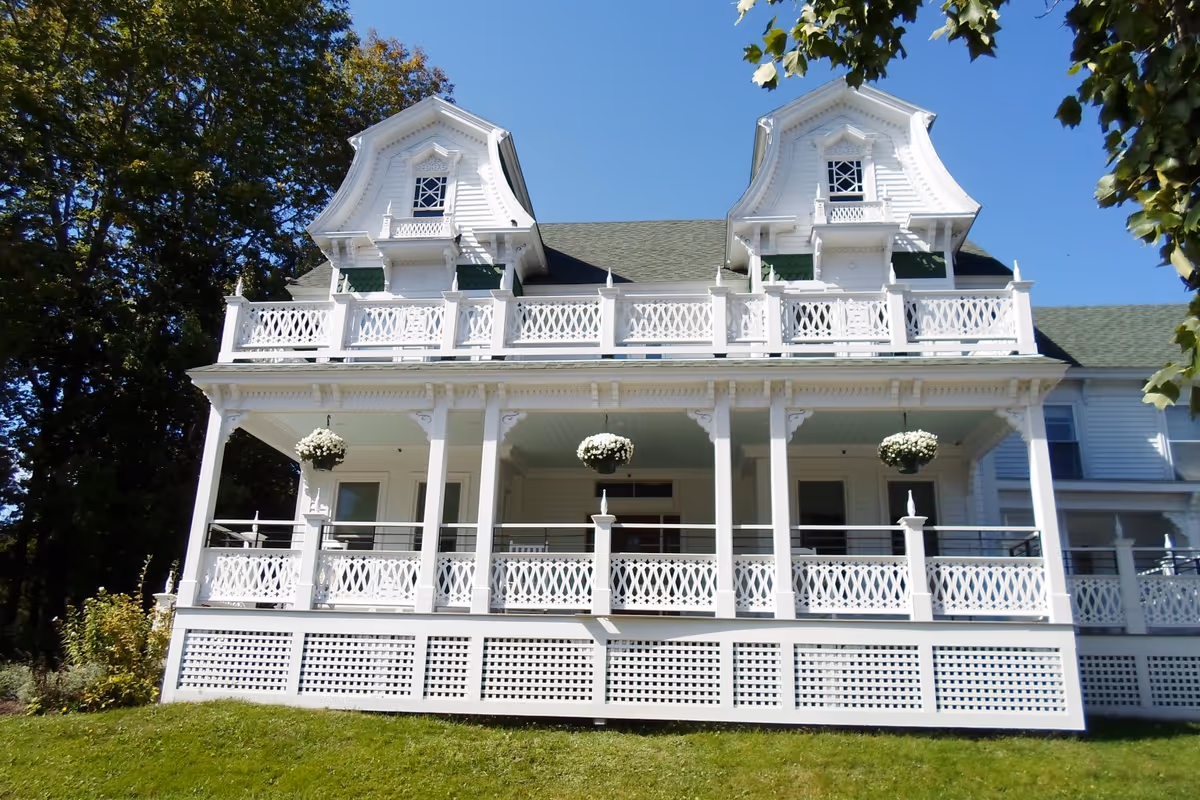 Front exterior view of a large white Victorian-style building with ornate railings and two dormer windows on the roof. The building has a covered porch with hanging flower baskets and is surrounded by green grass and trees under a clear blue sky.