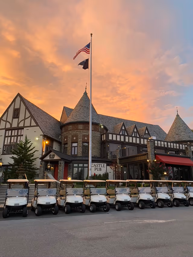 Tudor-style building with a flagpole and a row of parked golf carts in front under a colorful sunset sky.