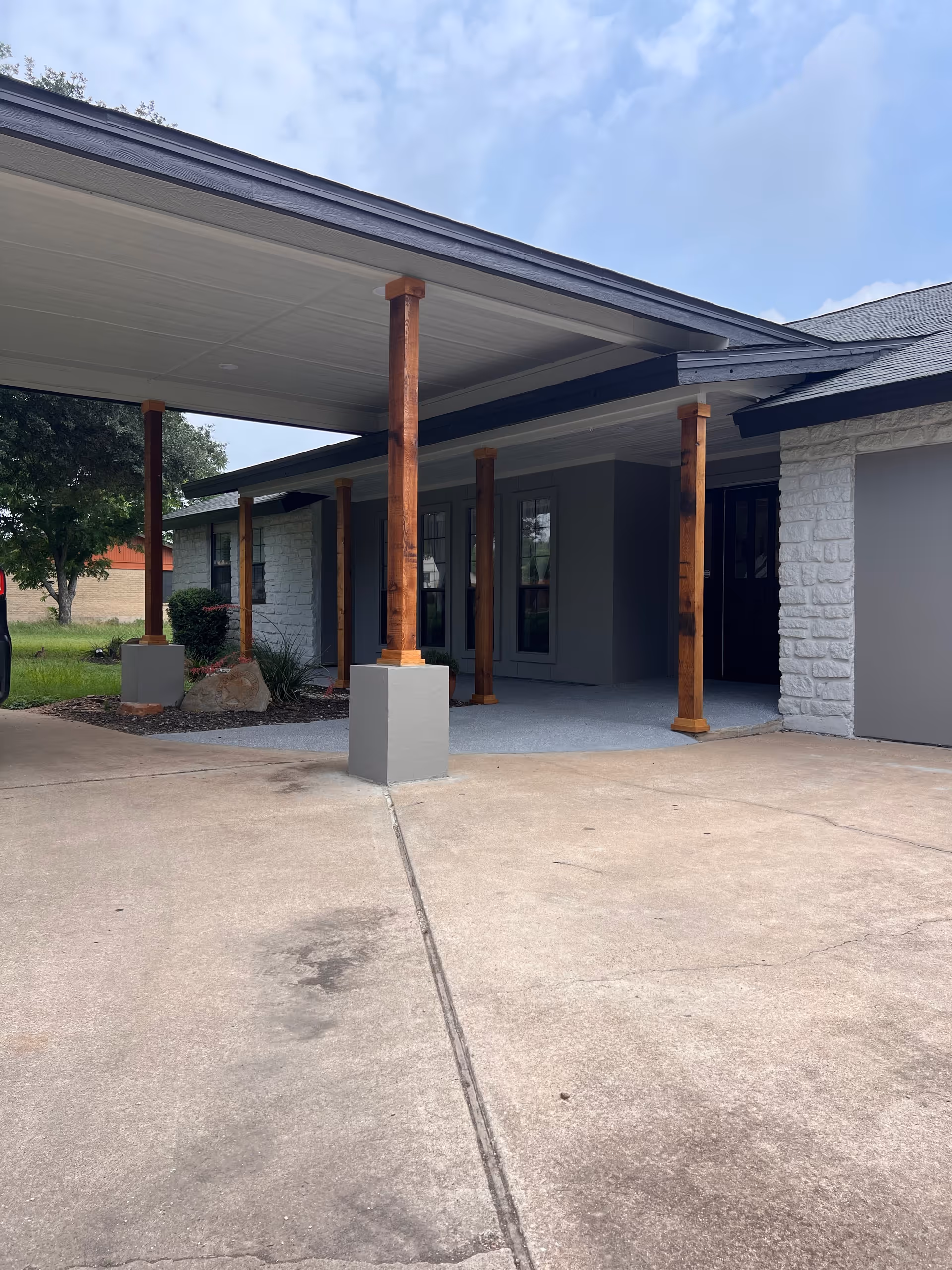 Exterior view of a senior living facility entrance with a covered porch supported by wooden pillars. The building has a stone and gray painted facade, with a concrete driveway in front and some greenery visible to the side under a partly cloudy sky.